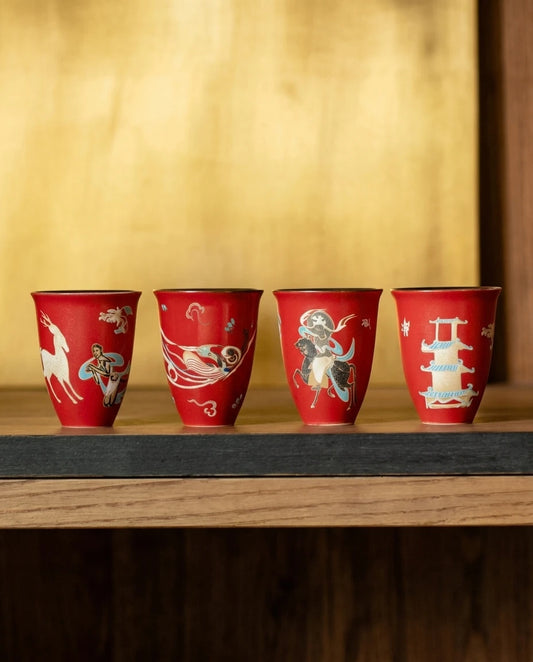 A set of four red ceramic Dunhuang tea cups with unique motifs on a wooden shelf